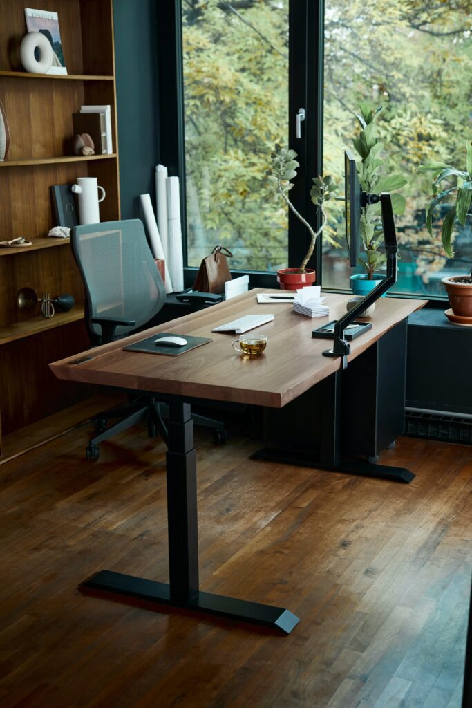 Modern home office with adjustable standing desk, ergonomic chair, computer setup, and indoor plants by a large window
